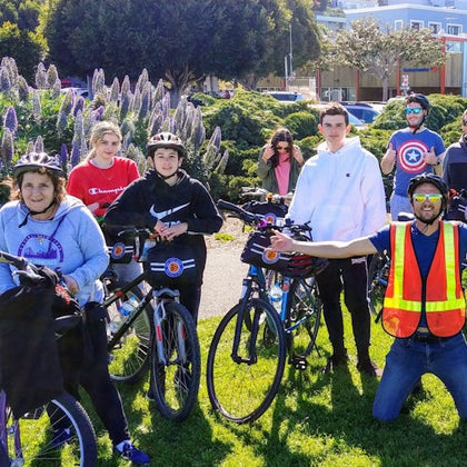 A group of people with bicycles, some wearing helmets and bright clothing, gathered outdoors near blooming flowers and greenery.