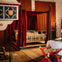 A medieval-style bedroom with a four-poster bed draped in red curtains, a decorated canopy, a fireplace, and a white table with red chairs.