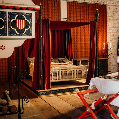 A medieval-style bedroom with a four-poster bed draped in red curtains, a decorated canopy, a fireplace, and a white table with red chairs.