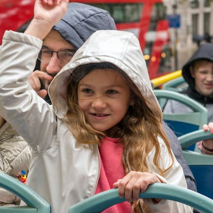 A young girl in a hooded jacket smiling and raising her arm, seated on an open-top bus with other passengers.