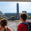 Two people on a boat observe a riverside view of the Tate Modern and the Millennium Bridge in London on a sunny day.