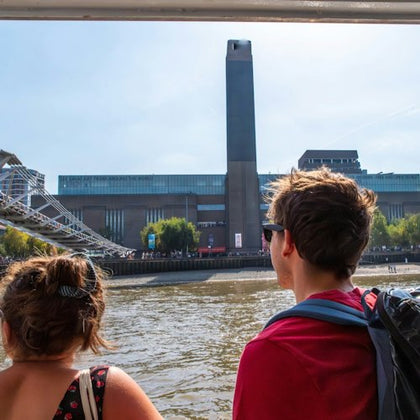 Two people on a boat observe a riverside view of the Tate Modern and the Millennium Bridge in London on a sunny day.