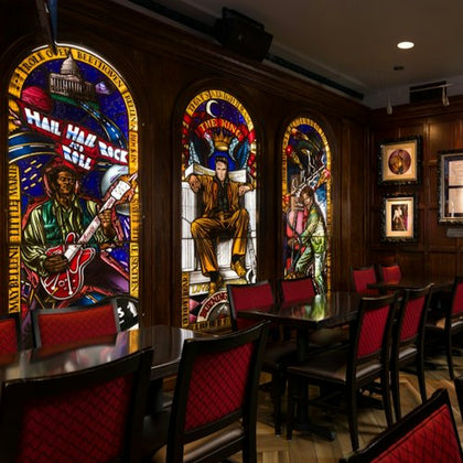 A room with stained glass art depicting musicians, framed pictures on walls, and tables with chairs featuring red upholstery.