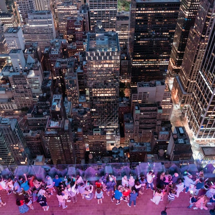 A rooftop observation deck with people overlooking a city skyline filled with tall buildings and illuminated windows at dusk.