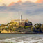A distant view of an island with a historic building and lighthouse, surrounded by water, under a colorful sky.