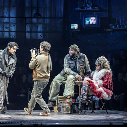 A theatrical performance featuring four actors on a dimly-lit stage, with props and a seated audience in the background.