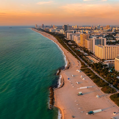 Aerial view of a coastline with beige sandy beach, turquoise water, and numerous high-rise buildings under a partly cloudy sky.