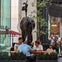 A group of people seated outdoors at a table near a large elephant sculpture in a city setting with trees and buildings.