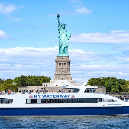 The Statue of Liberty with a NY Waterway ferry passing in the foreground on a sunny day.