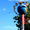 A signpost with a globe and various sculptures, including a coffee cup and pretzel, against a blue sky and tree background.