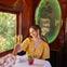 A woman in a yellow dress toasts with another person in a train dining car with a green-lit oval mirror.