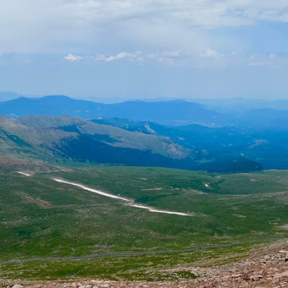 A scenic view of a grassy mountainous landscape with a winding trail, distant hills, and a partially cloudy sky.