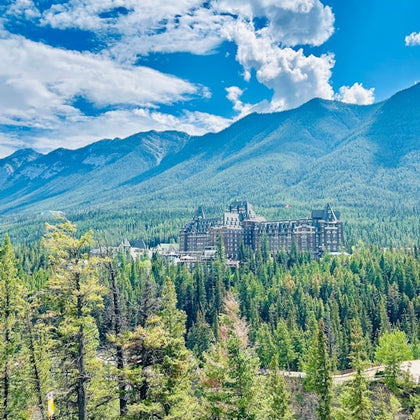 A large castle-like building surrounded by dense pine trees with mountains and a blue sky with clouds in the background.