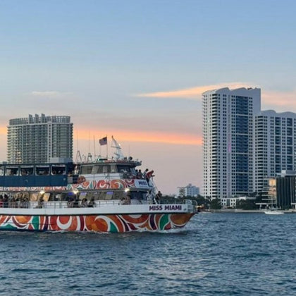 A colorful boat named "Miss Miami" on water with tall buildings and a sunset sky in the background.