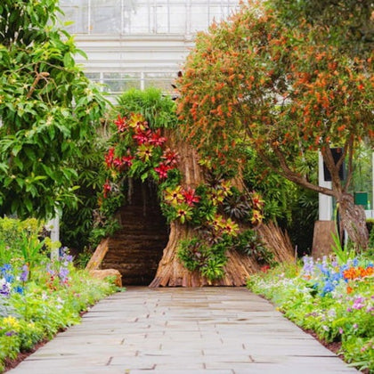 A garden path leading to a hollow tree trunk adorned with colorful flowers, surrounded by lush greenery and blooming plants.