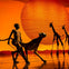 Performers with animal puppets on stage against a vivid orange background with a large circular element resembling the sun.