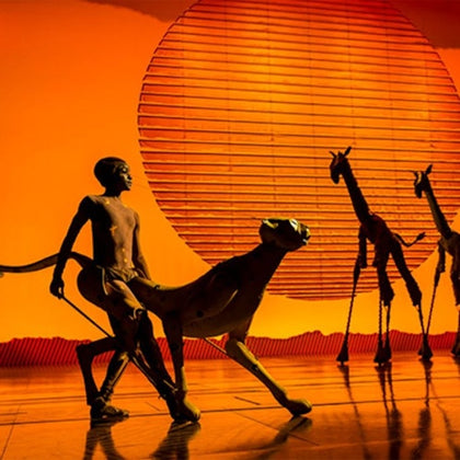 Performers with animal puppets on stage against a vivid orange background with a large circular element resembling the sun.