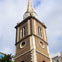 A tall brick clock tower with a spire, featuring arched windows and clocks on its sides, set against a cloudy sky.