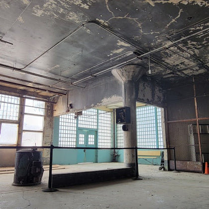 An empty industrial interior with large windows, exposed pipes, a concrete pillar, and peeling paint on walls and ceiling.