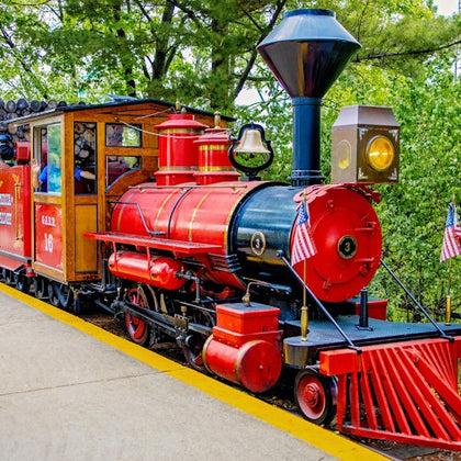 A red and black vintage-style steam locomotive on a railway platform, adorned with small American flags, surrounded by trees.