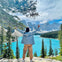 Person with raised arms stands before a turquoise lake, surrounded by pine trees and mountains under a partly cloudy sky.