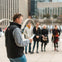 A man reads from a paper in an outdoor urban plaza, with several people standing in the background.
