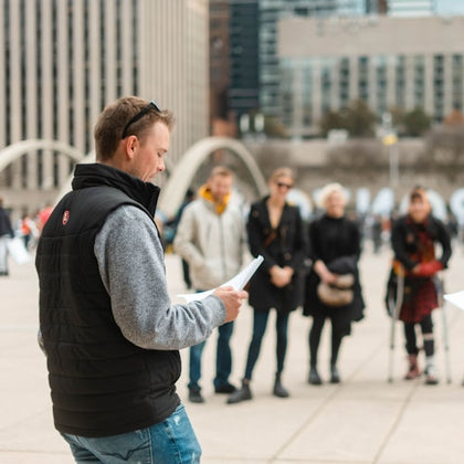 A man reads from a paper in an outdoor urban plaza, with several people standing in the background.