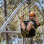 A person wearing a helmet rides a zip line through a wooded area, secured to the harness and smiling.