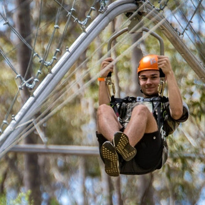 A person wearing a helmet rides a zip line through a wooded area, secured to the harness and smiling.