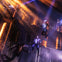 Performers in suits suspended in mid-air on a theatrical stage with dramatic lighting and industrial set design.