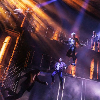 Performers in suits suspended in mid-air on a theatrical stage with dramatic lighting and industrial set design.