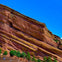 A large red rock formation with green trees at its base under a clear blue sky. Small figures are visible in the lower area.