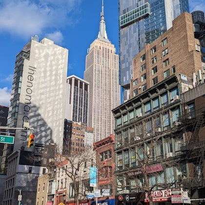A city street scene with buildings, including the Empire State Building, under a blue sky with scattered clouds.