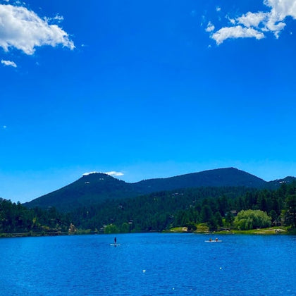 A serene lake with people on paddleboards, surrounded by forested hills under a blue sky with a few clouds.