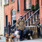 Person in a coat walking six dogs of various breeds on leashes in a residential neighborhood with brick and beige houses.