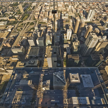 View from a glass observation deck high above a cityscape featuring numerous buildings, roads, and a distant highway.