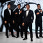 Four men in tuxedos stand together with arms crossed or hands on their chests, against a black and white backdrop.