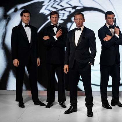Four men in tuxedos stand together with arms crossed or hands on their chests, against a black and white backdrop.