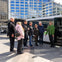 A group of people standing near a parked bus in an urban setting with modern buildings visible in the background.