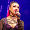 A performer on stage singing into a microphone, wearing a studded outfit and a choker, with a vibrant purple-lit background.