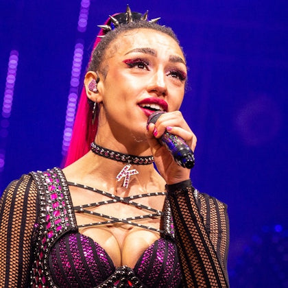 A performer on stage singing into a microphone, wearing a studded outfit and a choker, with a vibrant purple-lit background.