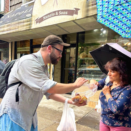A man hands food to a woman holding an umbrella outside a shop named Nunzio & Sons.