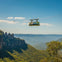 A scenic cable car suspended over a lush forest valley with rock formations and a clear blue sky in the background.