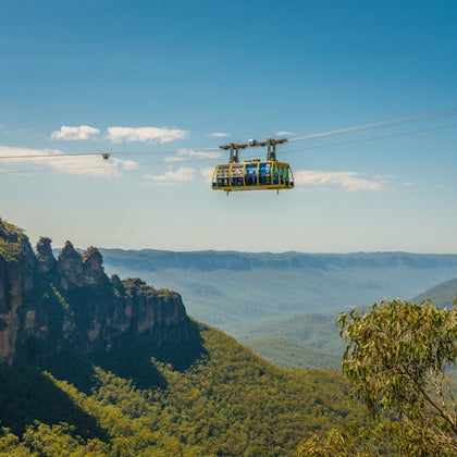 A scenic cable car suspended over a lush forest valley with rock formations and a clear blue sky in the background.