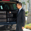 A man in a suit loads black suitcases into the back of a vehicle in an outdoor setting with palm trees and a building in the background.