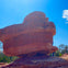 A large, reddish-brown rock formation under a clear blue sky, with a person partially visible near its base.