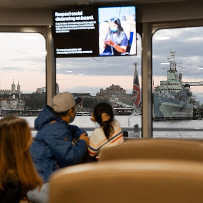 People sit on a ferry with a view of a warship and cityscape through the window. A screen displays a social distancing message.
