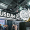 People in a museum lobby with a large coin sculpture and "Museum of" signage visible.