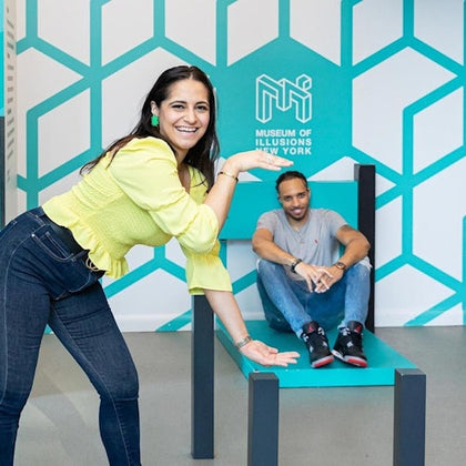 Two people at the Museum of Illusions in New York, interacting with an oversized chair exhibit creating a visual illusion.
