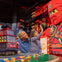 Child high-fives a person in a red LEGO ninja costume while an adult watches in a colorful LEGO-themed play area.